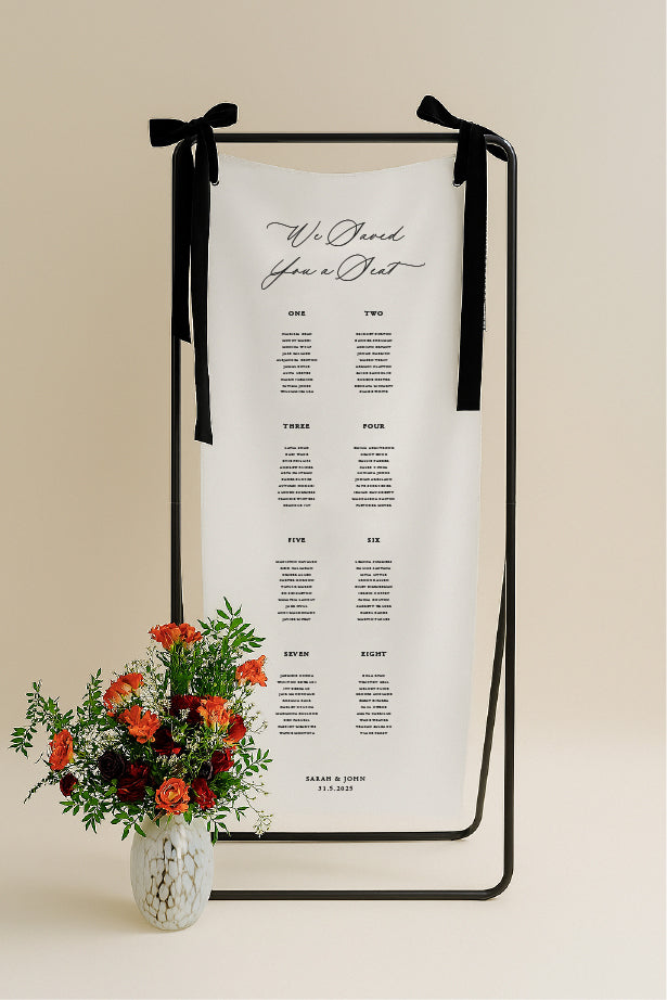 Decorative table seating chart with black ribbons and a vase of flowers on a beige background