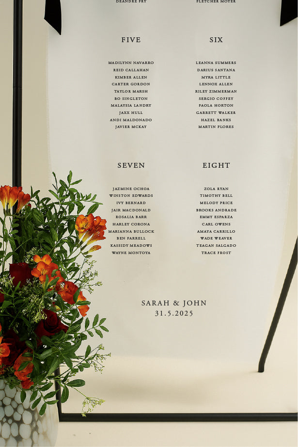 Framed seating chart with names and dates, accompanied by a vase of flowers.