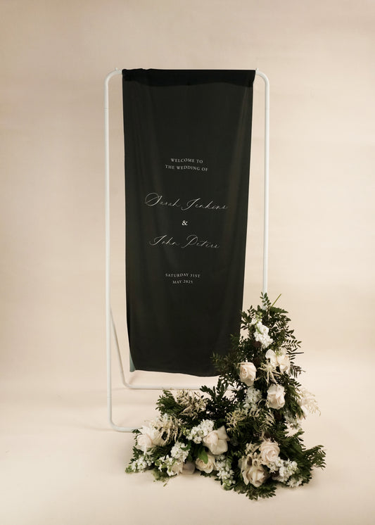 Black silk welcome sign with white text on a stand next to a floral arrangement
