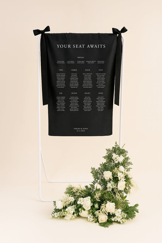 Black seating chart on a stand with floral arrangement on a beige background