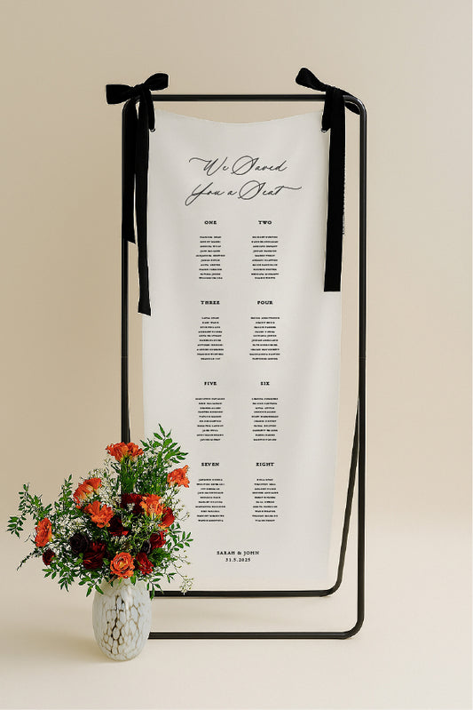 Decorative table seating chart with black ribbons and a vase of flowers on a beige background