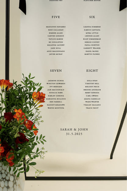 Framed seating chart with names and dates, accompanied by a vase of flowers.