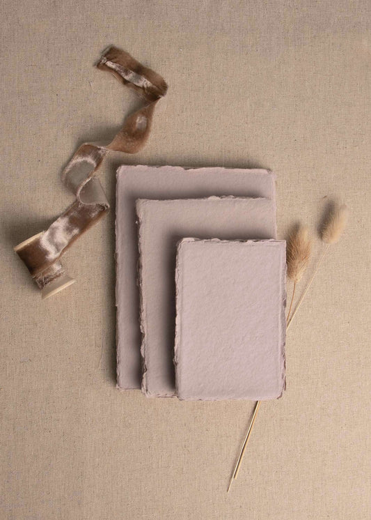 Pile of various sizes of Mauve handmade paper with deckle edge surrounded by dried flowers and velvet mocha ribbon spool
